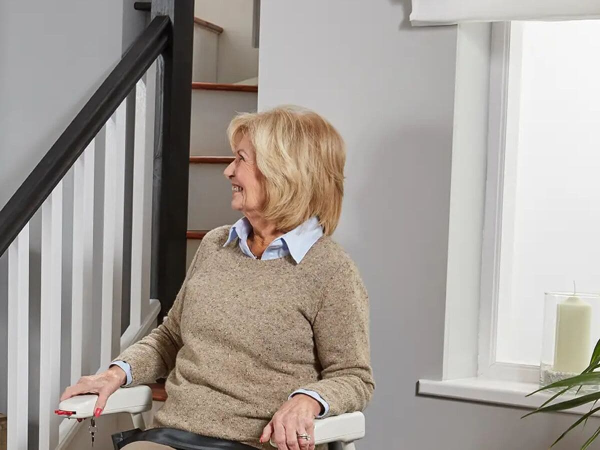 Une femme souriante assise sur un monte-escalier, au pied d’un escalier